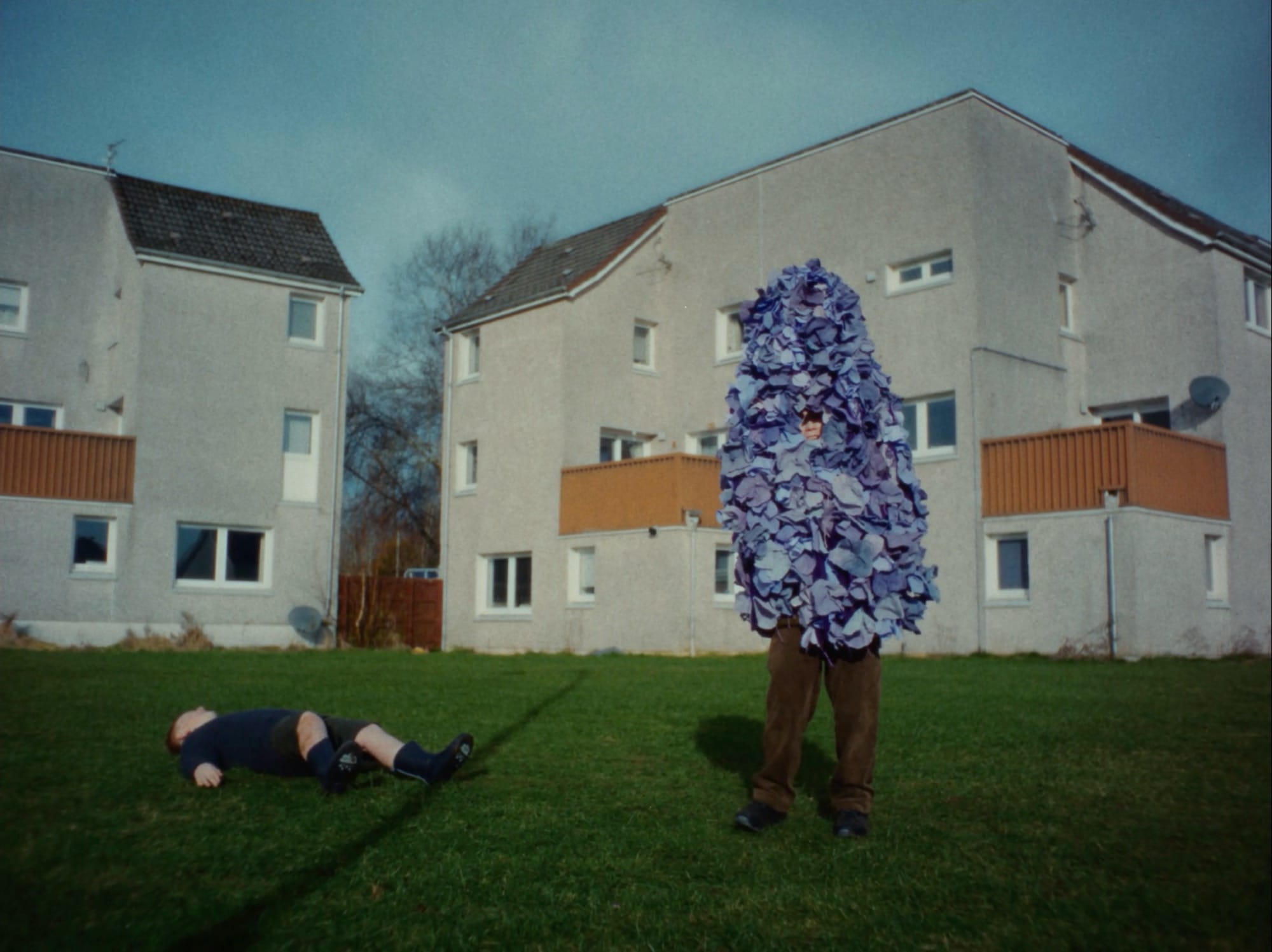a still from a video titled "A Ffern Fairytail" featuring children playing outside on a Scottish estate, one fallen to the ground and the other dressed like a giant purple "flower thing"